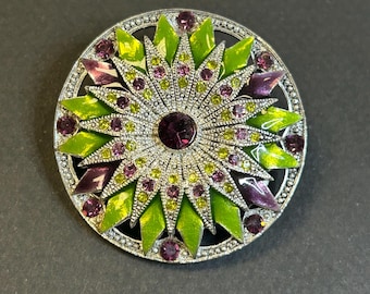 May include: A round, silver-toned brooch with a floral design. The brooch features a central purple gemstone surrounded by green, purple, and yellow enamel petals, and small clear crystals. The brooch has a textured border.