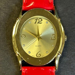 May include: A gold-tone watch with a red leather band. The watch face is oval-shaped and has a gold-tone bezel. The watch face is gold with black Roman numeral hour markers and black hands. The watch face also has a small gold star in the center and the word "QUARTZ" printed in black. The watch face also has the words "JAPAN" and "MOV'T" printed in black.