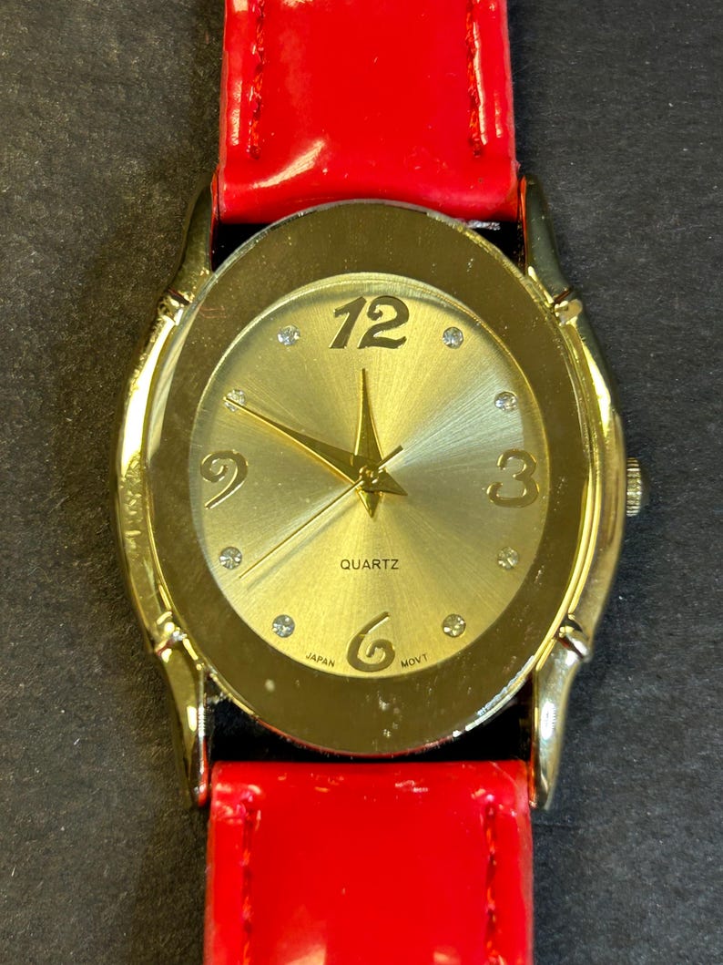 May include: A gold-tone watch with a red leather band. The watch face is oval-shaped and has a gold-tone bezel. The watch face is gold with black Roman numeral hour markers and black hands. The watch face also has a small gold star in the center and the word "QUARTZ" printed in black. The watch face also has the words "JAPAN" and "MOV'T" printed in black.