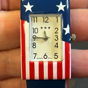 May include: A rectangular quartz watch with a red, white, and blue design. The watch face has a white background with black numerals and three stars. The watch band is blue, and the watch case features stars and stripes.