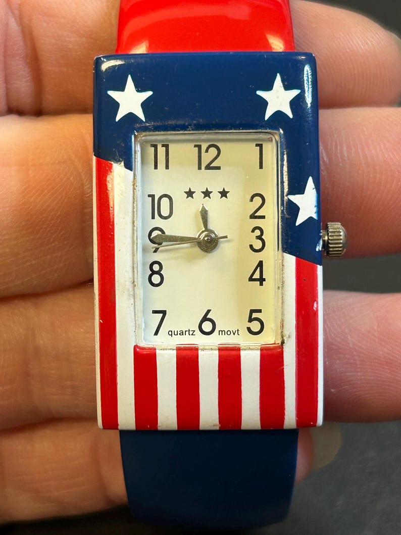 May include: A rectangular quartz watch with a red, white, and blue design. The watch face has a white background with black numerals and three stars. The watch band is blue, and the watch case features stars and stripes.