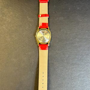 May include: A gold-tone wristwatch with a red leather band. The watch face is round and has a white background with black markings. The back of the watch has the words "Stainless Steel Back" and "Japan Movement" printed on it.