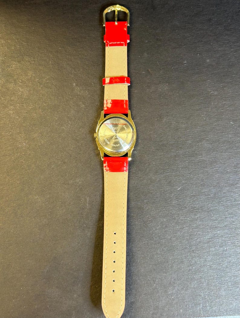 May include: A gold-tone wristwatch with a red leather band. The watch face is round and has a white background with black markings. The back of the watch has the words "Stainless Steel Back" and "Japan Movement" printed on it.