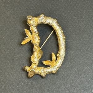 May include: Gold-toned brooch shaped like the letter D, formed from a stylised twig design with leaves.