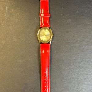 May include: A gold-tone wristwatch with a white face and black Roman numeral hour markers. The watch has a red leather band.