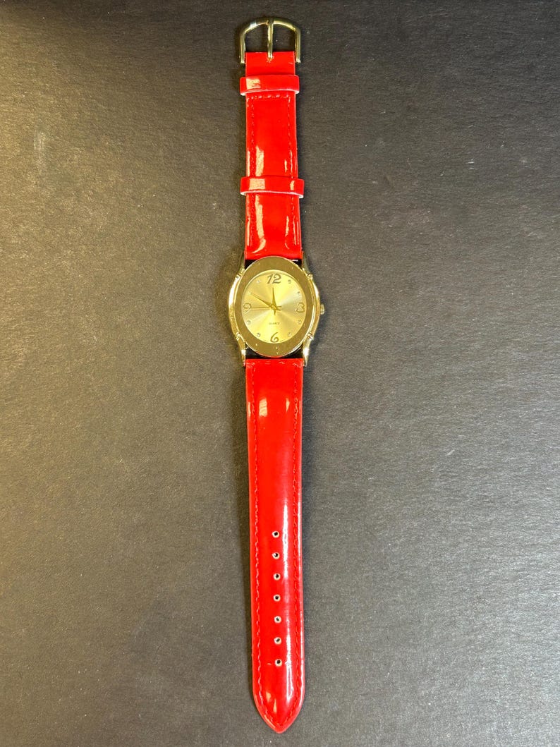 May include: A gold-tone wristwatch with a white face and black Roman numeral hour markers. The watch has a red leather band.