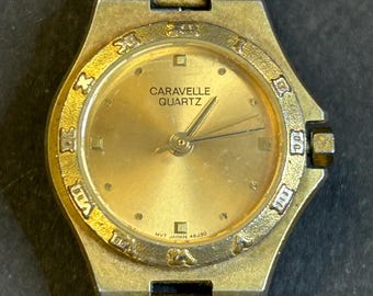 May include: A gold-toned Caravel quartz wristwatch with a gold-colored face and black hands. The watch face has a square at the 12 o'clock position and a small circle at the 4 o'clock position. The watch band is a gold-toned metal bracelet.