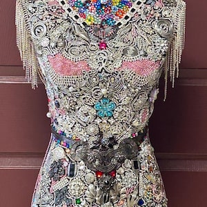 May include: A decorative dress form covered in an intricate array of embellishments. The form features a black base and is adorned with colorful beads, rhinestones, and silver-toned metalwork. Silver fringe hangs from the shoulders and hem.