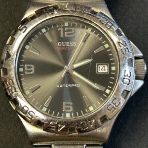 May include: A silver Guess watch with a black face and silver hands. The watch has a date window at the 3 o'clock position and says "WATERPRO" and "100M/330FT" on the face.