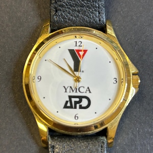 May include: A gold-tone wristwatch with a white face and black leather band. The face features a red and black YMCA logo and the text "YMCA APD".