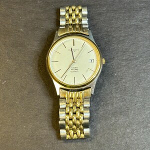 May include: A gold and silver two-tone watch with a white face. The watch face has black Roman numeral hour markers and a date window at the 3 o'clock position. The watch band is a metal link band. The watch face says "DuMont&eacute;" and "Lucien Piccard Quartz".