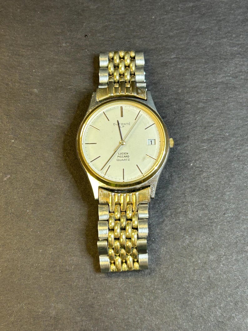 May include: A gold and silver two-tone watch with a white face. The watch face has black Roman numeral hour markers and a date window at the 3 o'clock position. The watch band is a metal link band. The watch face says "DuMont&eacute;" and "Lucien Piccard Quartz".