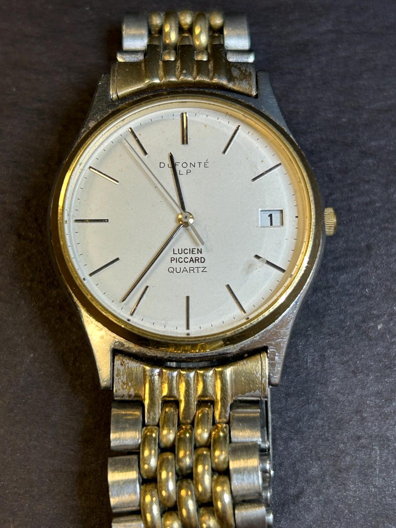 May include: A two-tone gold and silver watch with a white face. The watch face has black hands and black Roman numeral hour markers. The watch face also has the text "DuPont&eacute; LP", "Lucien Piccard", and "Quartz".