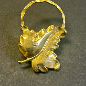 Pode incluir: Broche dourado em forma de folha com uma pega texturizada. O broche apresenta veios detalhados na folha e um acabamento liso e polido. O design é reminiscente da natureza e de temas botânicos.