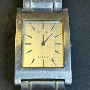 May include: A rectangular Guess watch with a silver-tone metal strap and case. The watch face is gold with black hands and markers. The word "GUESS" is printed at the top, and "STEEL" is printed at the bottom.