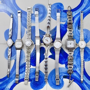 Peut inclure: Une collection de montres-bracelets argent&eacute;es vintage avec diff&eacute;rents styles de bracelets, dont des mailles et des motifs orn&eacute;s. Les cadrans des montres sont principalement blancs avec des chiffres et des aiguilles noirs. Les montres sont pr&eacute;sent&eacute;es sur une surface &agrave; motifs bleus et blancs.