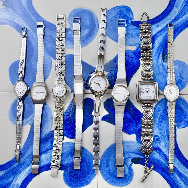 Peut inclure: Une collection de montres-bracelets argent&eacute;es vintage avec diff&eacute;rents styles de bracelets, dont des mailles et des motifs orn&eacute;s. Les cadrans des montres sont principalement blancs avec des chiffres et des aiguilles noirs. Les montres sont pr&eacute;sent&eacute;es sur une surface &agrave; motifs bleus et blancs.