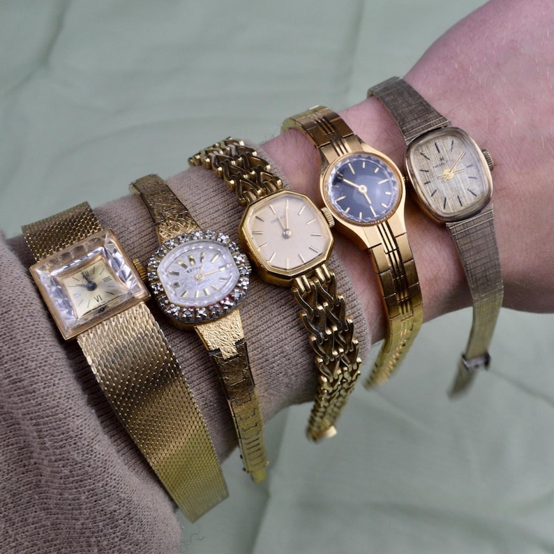 May include: A collection of five vintage gold-toned wristwatches, each with a unique design. The watches feature various band styles, including mesh, link, and textured bands. The watch faces showcase different shapes and colors, including a square, oval, and round face.