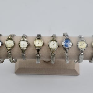 Puede incluir: Una colección de ocho relojes de pulsera vintage expuestos en un soporte beige. Los relojes presentan varias correas de metal plateado y esferas de reloj de diferentes formas, incluyendo redondas, cuadradas y ovaladas. Las esferas de los relojes tienen diferentes colores, incluyendo blanco y azul.