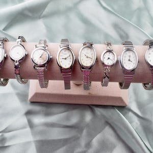 May include: Seven vintage wristwatches with silver-toned metal straps and white faces, displayed on a pink stand. The watch faces are various shapes, including round and oval. The watches are displayed on a pink stand.