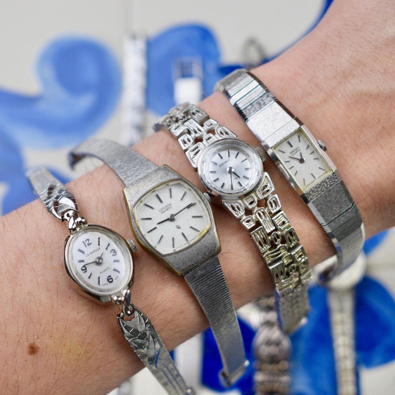 Peut inclure: Quatre montres-bracelets argent&eacute;es vintage avec des cadrans blancs sont pr&eacute;sent&eacute;es sur un poignet. Les montres ont diff&eacute;rents styles de bracelets, dont un bracelet &agrave; maillons, un bracelet en maille et un cadran rectangulaire. Les cadrans ont des index et des aiguilles noirs.