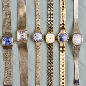 May include: A collection of six vintage gold-toned wristwatches. The watches feature various face shapes, including square, oval, and rectangular, with different band styles. Some have blue or cream-colored faces, and some are adorned with decorative elements.