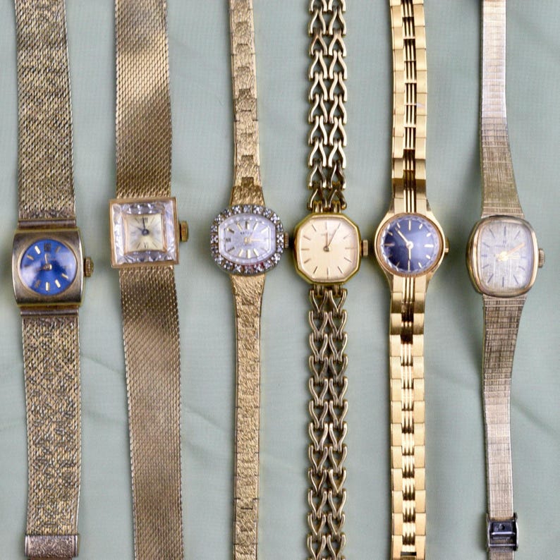 May include: A collection of six vintage gold-toned wristwatches. The watches feature various face shapes, including square, oval, and rectangular, with different band styles. Some have blue or cream-colored faces, and some are adorned with decorative elements.