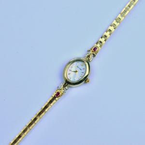May include: A gold-tone wristwatch with an oval-shaped case and a mother-of-pearl dial featuring the Waltham logo. The watch band is composed of gold-tone links, and the lugs are accented with small clear stones and a single red gemstone on each side.