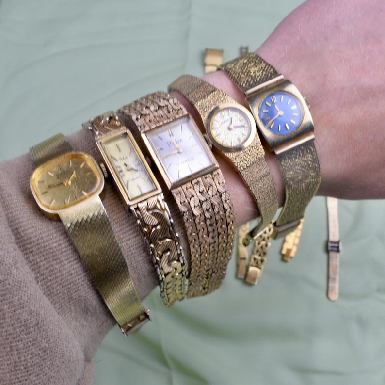 May include: A collection of vintage gold-toned wristwatches with various band styles, including mesh and linked designs. The watch faces are in different shapes, with some featuring Roman numerals and a blue face.