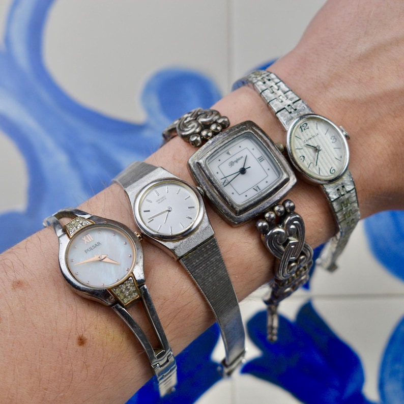 Peut inclure: Quatre montres-bracelets argent&eacute;es avec des cadrans blancs. L'une a un cadran en nacre et des d&eacute;tails en cristal. Une autre a un cadran carr&eacute; avec des chiffres romains. Les montres sont pr&eacute;sent&eacute;es sur un poignet.