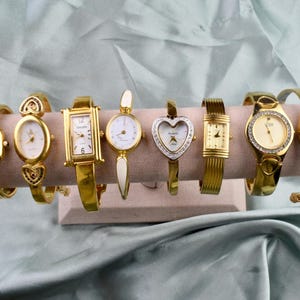 Pode incluir: Uma coleção de oito relógios de pulso dourados exibidos em um suporte castanho claro. Os relógios apresentam várias formas de mostrador, incluindo designs quadrados, ovais e em forma de coração. Alguns relógios são adornados com strass.