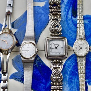 Peut inclure: Plusieurs montres-bracelets vintage avec des bracelets en m&eacute;tal argent&eacute; et des cadrans blancs. L'une des montres a un cadran en nacre et une lunette en forme de diamant. Une autre montre a un cadran carr&eacute; et un bracelet d&eacute;coratif. Les montres sont expos&eacute;es sur une surface &agrave; motifs bleus et blancs.