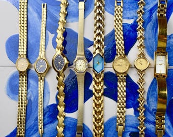May include: A collection of eight gold-toned wristwatches with various band styles and face shapes. The watch faces include round, oval, and rectangular designs, with some featuring gemstones and colored accents. The watches are displayed on a blue and white patterned surface.