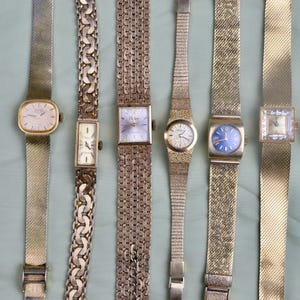 May include: A collection of vintage gold-toned wristwatches with various band styles, including mesh and link chains. The watch faces are in different shapes, with some featuring Roman numerals and others with a minimalist design. The watches are displayed on a light green surface.