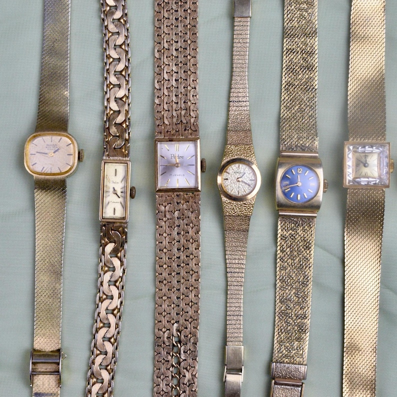 May include: A collection of vintage gold-toned wristwatches with various band styles, including mesh and link chains. The watch faces are in different shapes, with some featuring Roman numerals and others with a minimalist design. The watches are displayed on a light green surface.