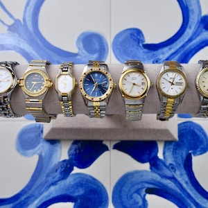 Può includere: Sette orologi da polso assortiti esposti su un supporto grigio. Gli orologi presentano vari design, con tonalità oro e argento, e quadranti di diversi colori. Alcuni hanno cinturini in metallo, altri in pelle. Lo sfondo è una piastrella a motivi blu e bianchi.
