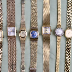 May include: A collection of vintage gold-toned wristwatches with various band styles and face designs. The watches feature square, oval, and round faces, some with intricate detailing and different colored dials, including blue and white.