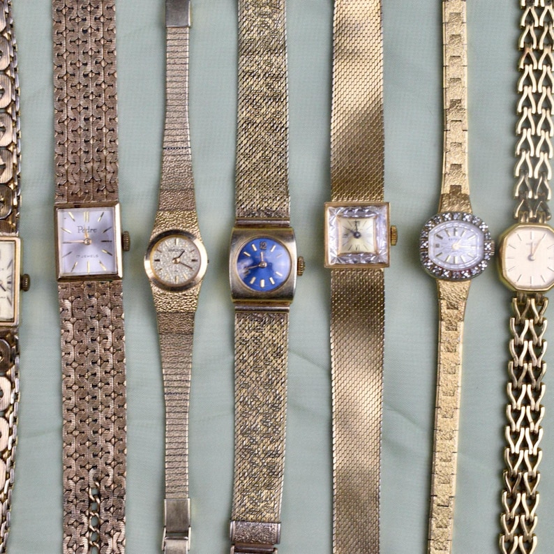 May include: A collection of vintage gold-toned wristwatches with various band styles and face designs. The watches feature square, oval, and round faces, some with intricate detailing and different colored dials, including blue and white.