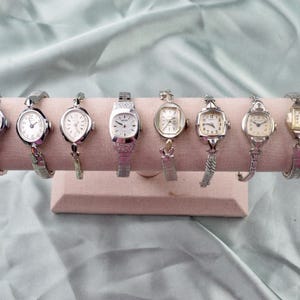 May include: Eight vintage wristwatches are displayed on a pink, cylindrical stand. The watches have silver-toned metal straps and watch faces in white, cream, and gold. The background is a light blue fabric.