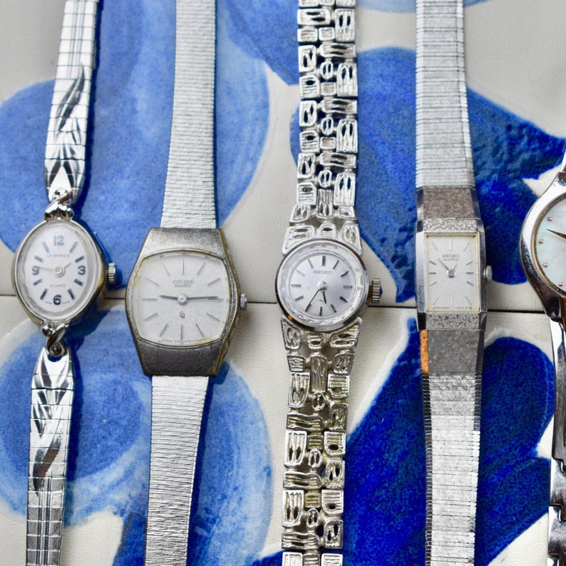 Peut inclure: Plusieurs montres-bracelets argent&eacute;es vintage avec diff&eacute;rents mod&egrave;les de bracelets et de cadrans. Les montres ont des cadrans blancs avec des chiffres et des aiguilles noirs. Les bracelets pr&eacute;sentent des motifs et des textures complexes. Le fond est un motif floral bleu et blanc.