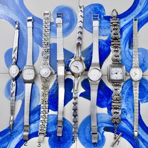 Peut inclure: Collection de huit montres-bracelets argent&eacute;es avec diff&eacute;rents designs de bracelets et cadrans blancs. Les montres sont pr&eacute;sent&eacute;es sur une surface &agrave; motifs bleus et blancs, pr&eacute;sentant une gamme de styles.