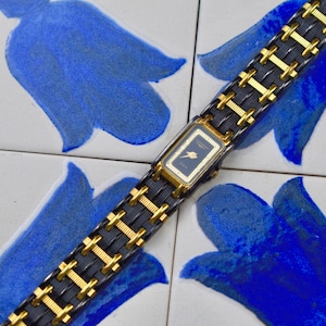 May include: A rectangular wristwatch with a dark face and gold-toned detailing. The watch features a black strap with gold-coloured links. The watch is set against a background of white tiles with blue floral designs.