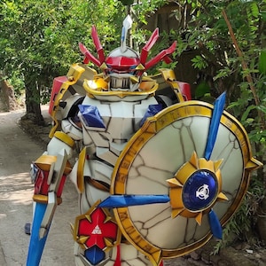 Peut inclure: Un costume de robot blanc, rouge, bleu et or avec un grand bouclier et une épée. Le bouclier a une bordure dorée et un centre bleu avec un symbole blanc. Le robot porte un casque avec une crête rouge.