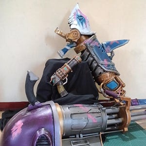 May include: A detailed, multi-colored cosplay prop resembling a futuristic weapon. The prop features a large, purple and gray barrel with multiple gun barrels, and a complex upper structure with blue, gold, and silver accents. The design includes intricate details and a stylized helmet.