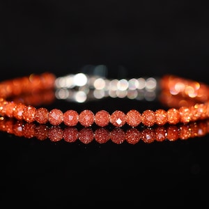 Puede incluir: Una pulsera de cuentas rojo-anaranjadas brillantes con un cierre plateado. Las cuentas son pequeñas y redondas, reflejando la luz. La pulsera se muestra sobre un fondo negro, con su reflejo visible.