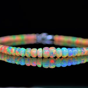 Könnte beinhalten: Ein farbenfrohes Opalperlenarmband mit einem silbernen Verschluss. Die Perlen zeigen eine Reihe von Farben, darunter Orange, Grün, Blau und Gelb. Das Armband spiegelt sich in der dunklen Oberfläche darunter wider.