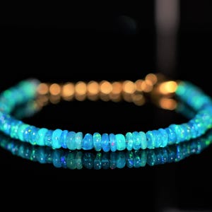 May include: A delicate bracelet featuring small, round, blue-green opal beads. The beads are strung together with a gold-colored clasp. The bracelet is displayed on a reflective black surface, creating a mirrored effect.