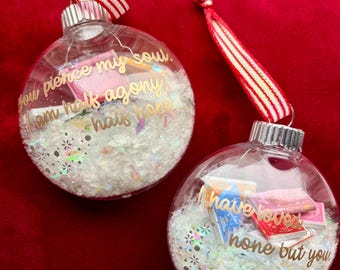 May include: Two clear Christmas baubles with gold text and red and white striped ribbons. The baubles contain miniature books and glitter. One reads "You pierce my soul, I am half agony, half hope." The other reads "I have loved none but you."