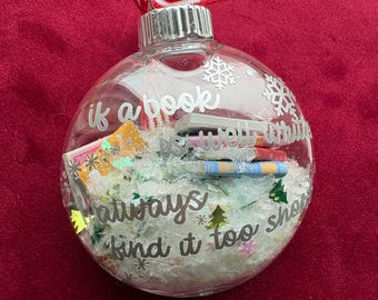 May include: Clear glass Christmas ornament with a silver cap and a red and white striped ribbon. The ornament contains miniature books and decorative accents. Text on the ornament reads "if a book sells well, I always find it too short."
