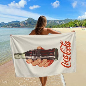May include: A white beach towel featuring a vintage Coca-Cola logo and a hand holding a bottle of Coca-Cola.